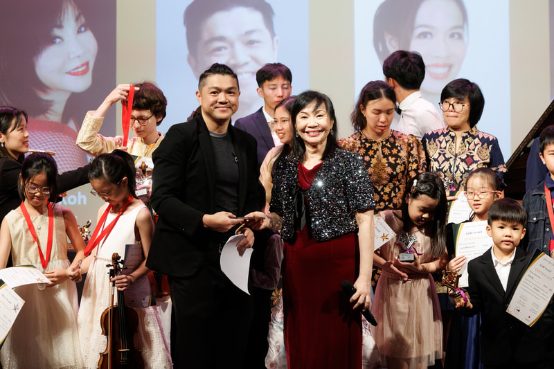 Group of children and adults standing on stage during an awards ceremony, with large portraits of two women and a man projected in the background.