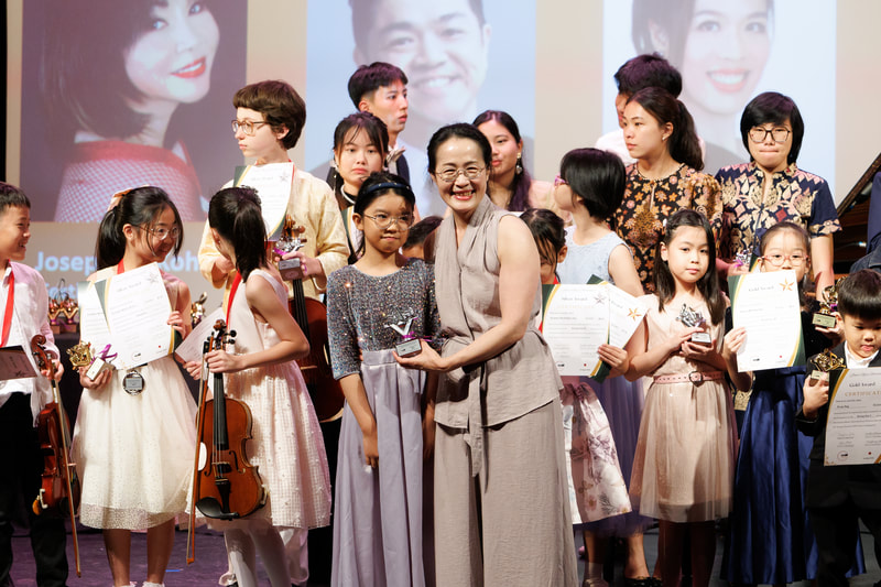 A group of children and adults on stage during an awards ceremony, with some holding certificates and trophies, and a woman in the center smiling holding a trophy.