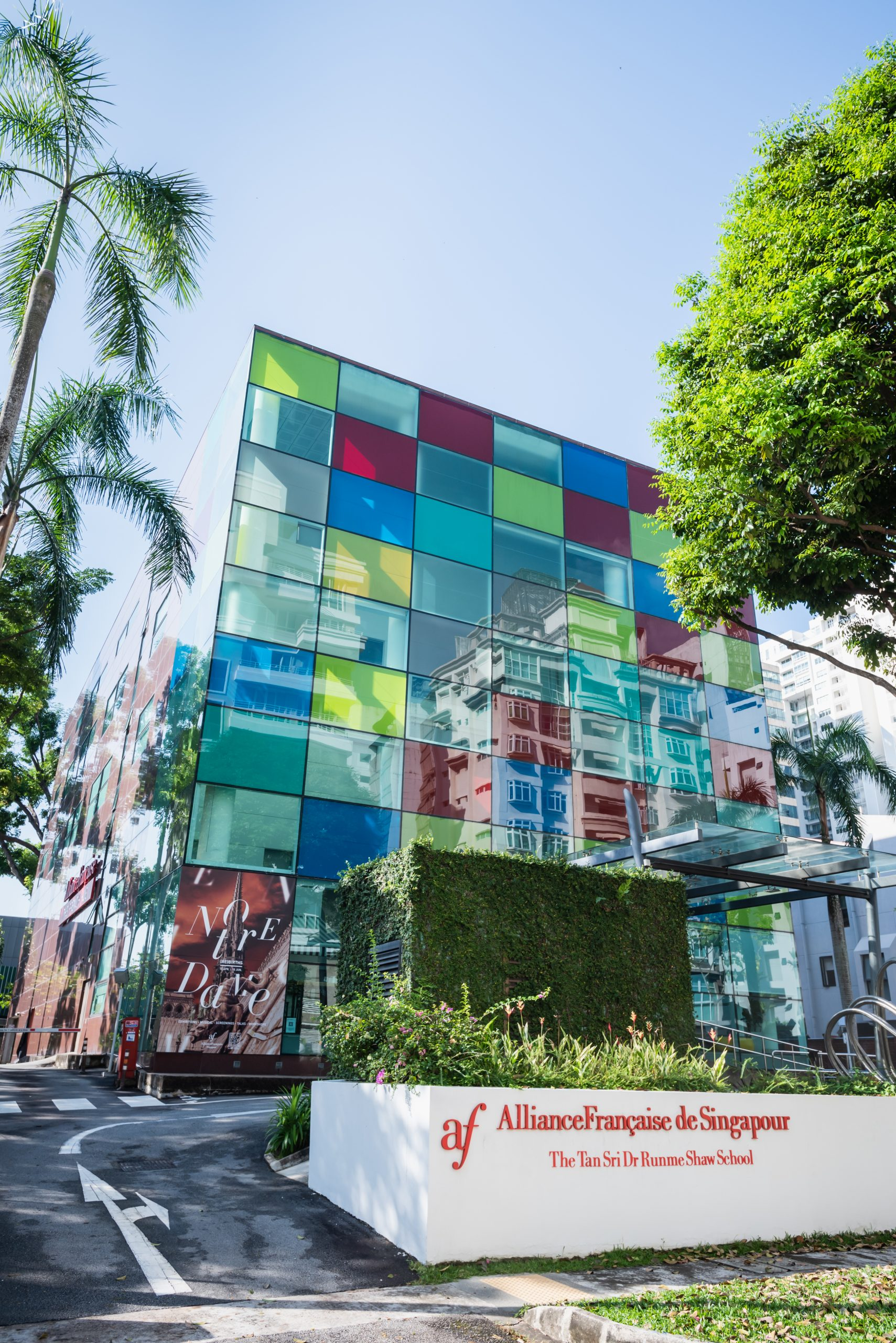 A modern glass building with colorful panels reflecting surrounding structures, and a sign in front reads 'Alliance Française de Singapour, The Tan Sri Dr Runme Shaw School' with a red logo, surrounded by lush greenery and palm trees.