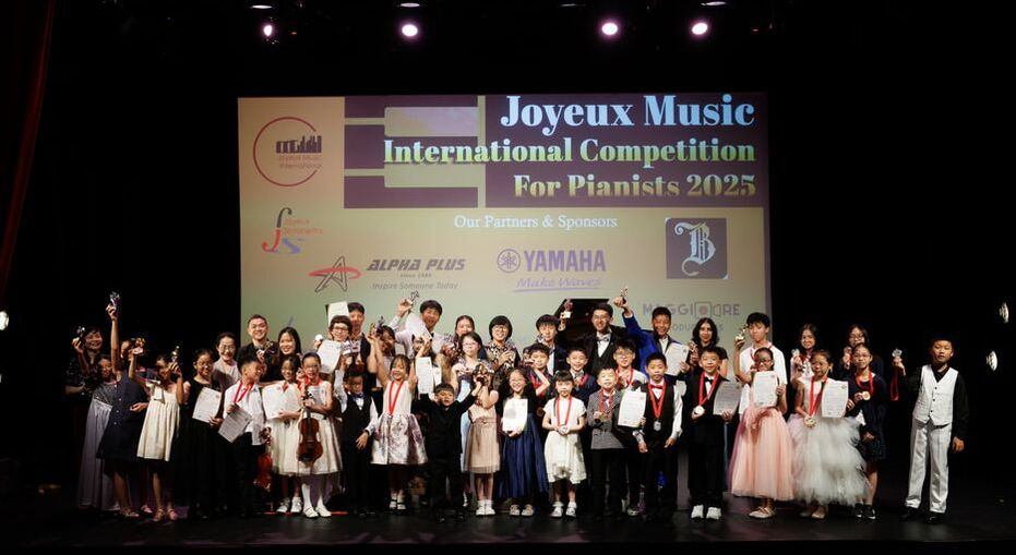 Group of children and adults on stage at the Joyeux Music International Competition for Pianists 2025, holding certificates and medals, with a large screen behind them displaying event information and sponsors.