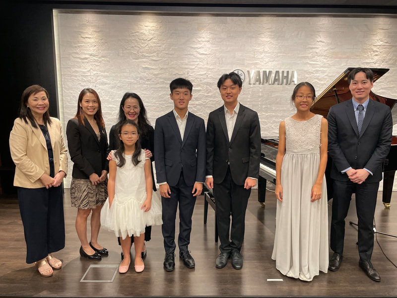 A group of eight people, including children and adults, standing on a stage in front of a white wall with a Yamaha logo and a grand piano, at a music recital or concert.