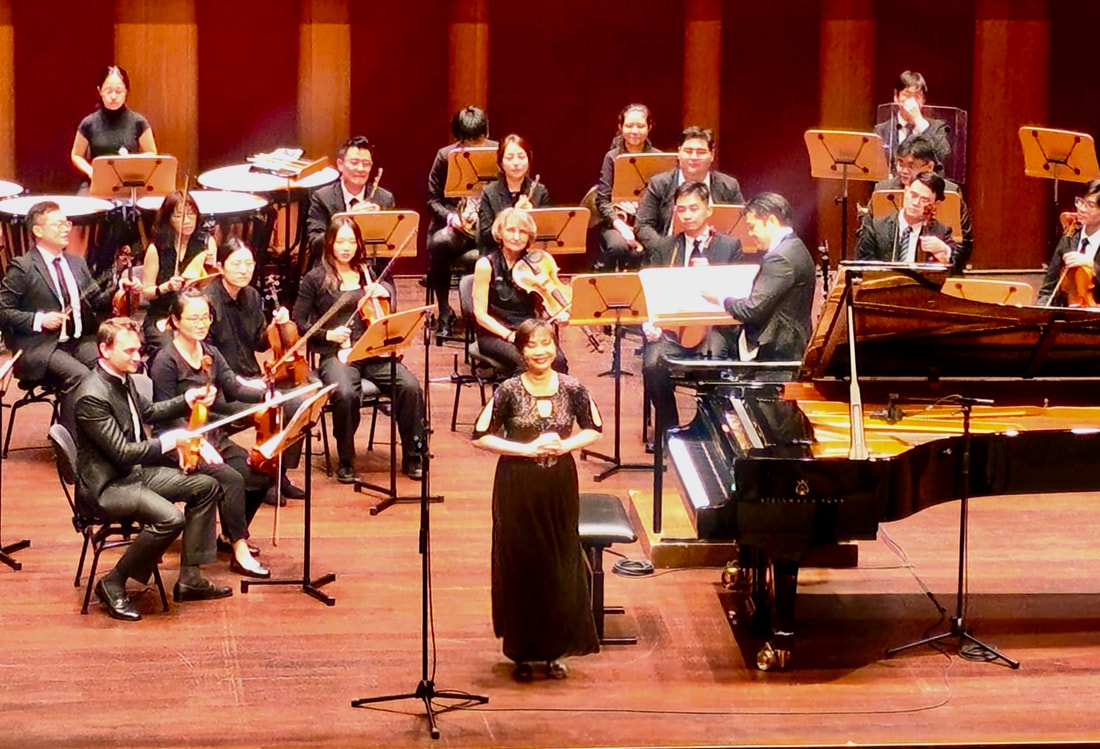 Orchestra performing on stage with a woman standing in front, holding a microphone, next to a grand piano.