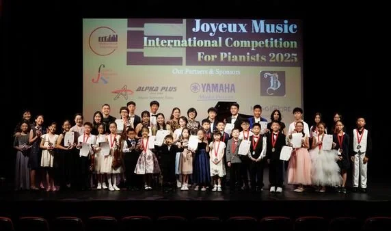 Group of children on stage holding certificates and medals at the Joyeux Music International Competition for Pianists 2025, with a large screen behind them displaying event details and sponsor logos.
