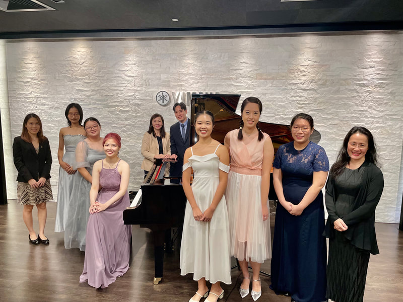 A group of nine people, mostly women, posing with a grand piano in a room with a white brick wall. They are dressed in formal or semi-formal attire, and some are smiling.