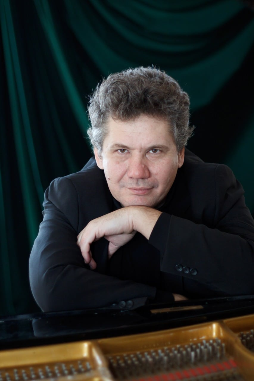 A man with curly hair and light skin, wearing a black jacket, leaning on a musical instrument with a green and black background.