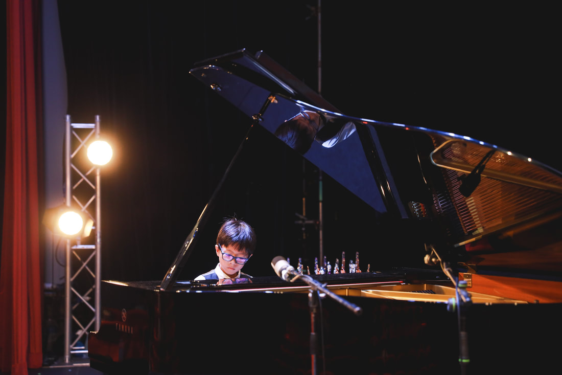 A young boy playing a grand piano on stage, with stage lights and a microphone nearby.