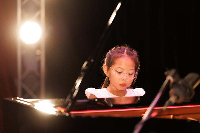 A young girl with glasses playing a grand piano on stage, focused with a serious expression.