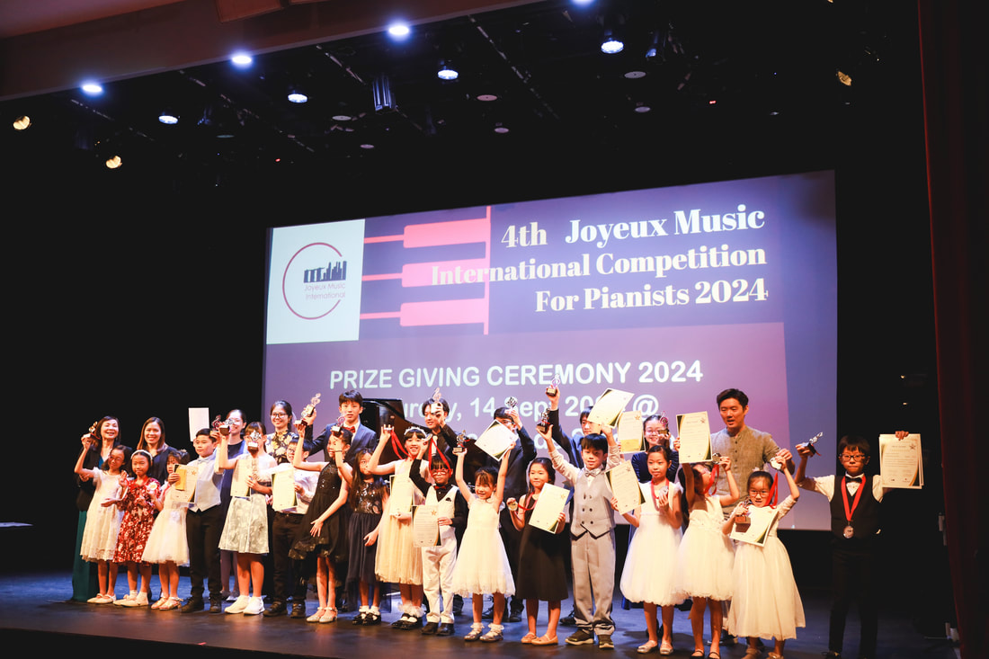 Group of children and adults on stage at a music competition award ceremony, holding certificates and trophies, with a large screen in background displaying event details.