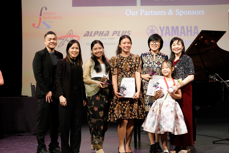Group of seven people, including six women and one young girl, standing on stage at an awards or recognition event, holding certificates and trophies, with a piano to the right, and logos of sponsors projected on the screen behind them.
