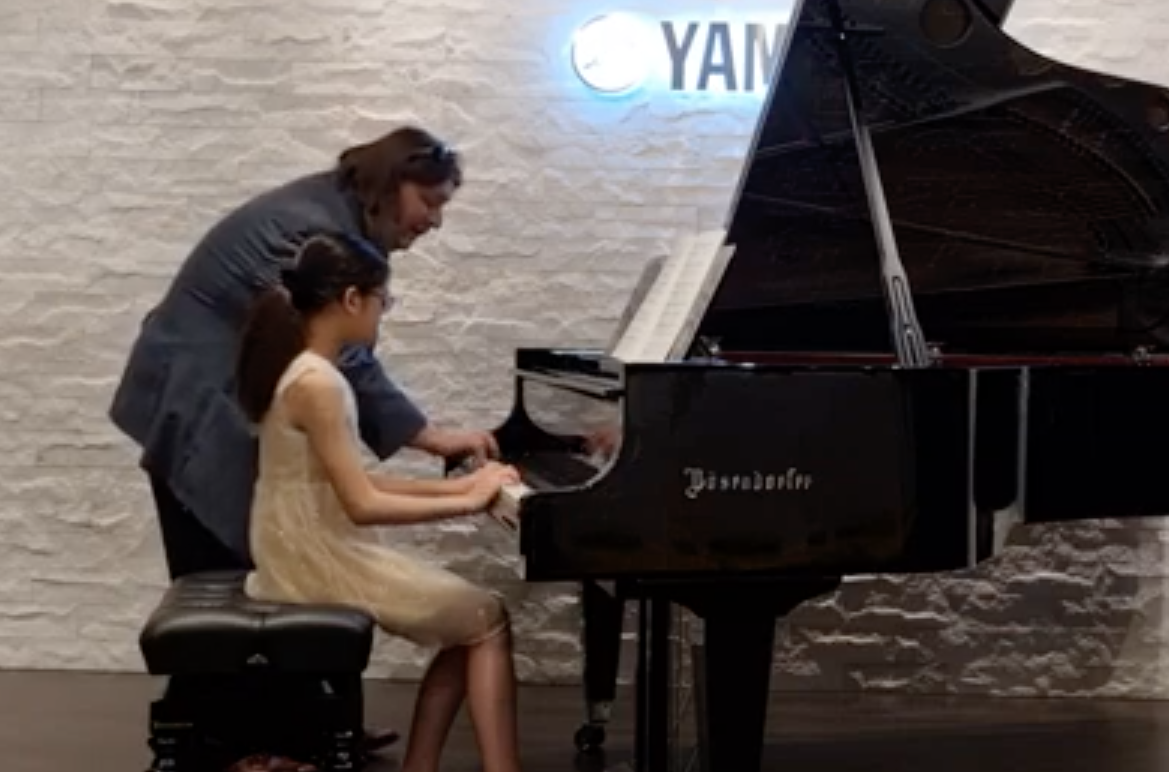 A young girl playing the piano with an adult guiding her, in a room with a white brick wall and Yamaha sign.