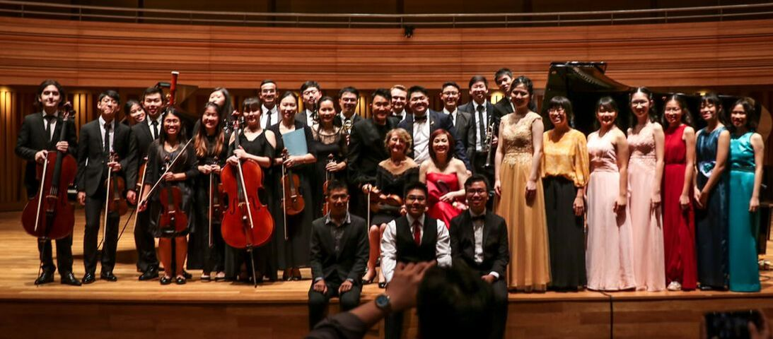 Group of musicians and singers on stage after a performance, some holding instruments, in formal attire, with a grand piano in the background.