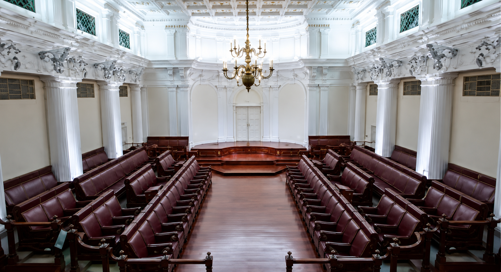 Elegant courtroom with white walls, tall columns, gold chandelier, and rows of red leather chairs arranged facing a wooden bench and a door in the background.