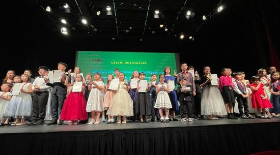 Group of children standing on stage holding certificates, dressed in formal and colorful clothing, in front of a large green screen that reads 'Our Mission.'