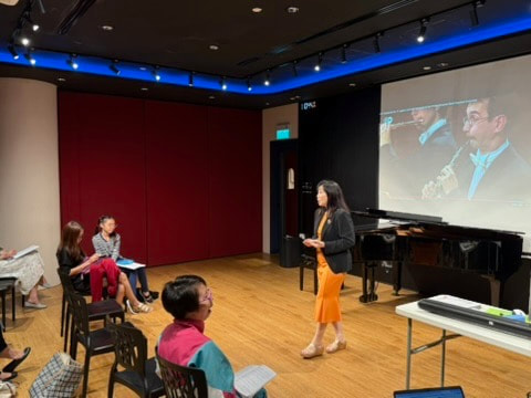 A woman presenting to a small audience in a room with a large screen displaying her image, a piano, and a table with equipment.