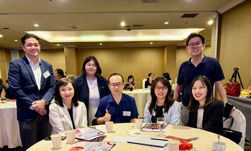 Group of seven people, four women and three men, gathered around a table at a conference or event in a banquet hall, smiling for a photo.