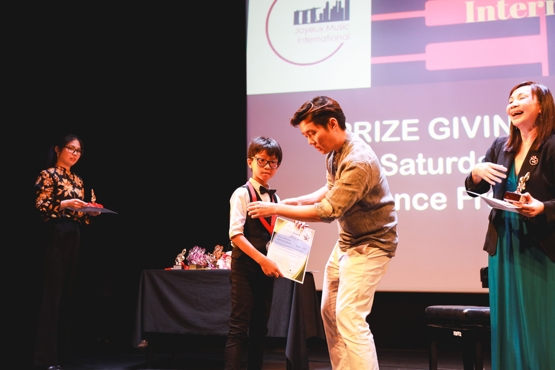 A young boy receiving a certificate and medal from a man during an award ceremony on stage. Two women, one holding a plaque, are also present on stage. A table behind them displays trophies and awards. A large screen in the background displays the event logo and text about prize giving.