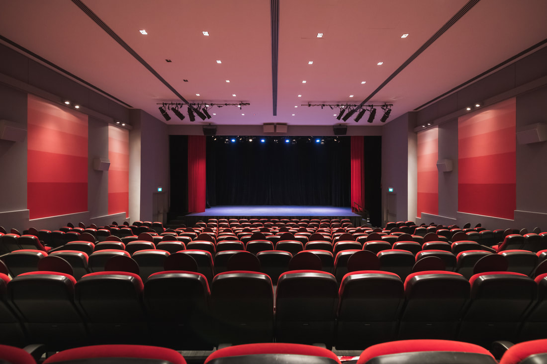 Empty theater showing a stage with black curtains and red side curtains, illuminated with stage lights, with rows of red and black seats.