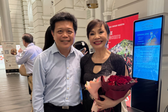 A man and woman smiling indoors at an event, with the woman holding a bouquet of red roses.