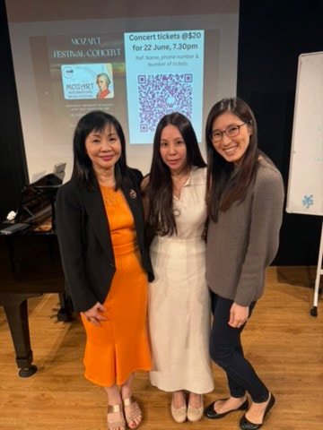 Three women standing together in a room with a piano and a projection screen behind them. The woman on the left is wearing a black blazer and orange dress, the woman in the middle is in a white dress, and the woman on the right is in a gray sweater and dark pants. They are smiling at the camera.