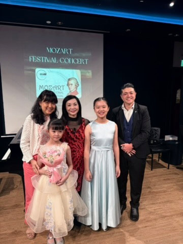 Five people, including two young girls and three adults, standing together in front of a presentation screen at the Mozart Festival concert, all smiling.