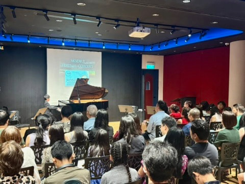 Audience attending a piano concert in a small auditorium, with a grand piano on stage and a presentation screen.