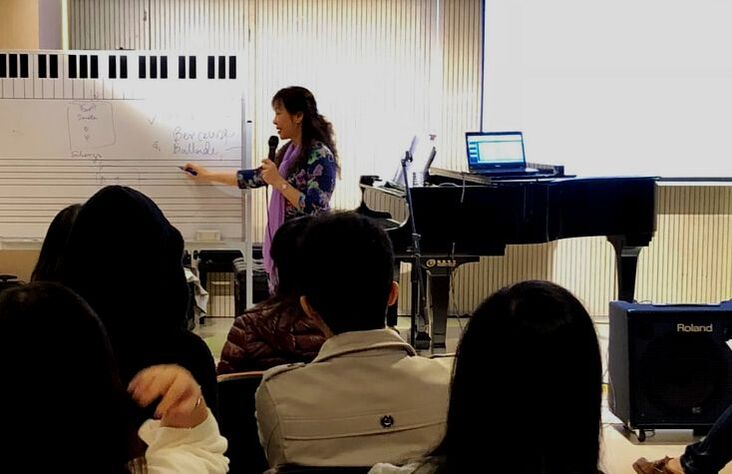 A woman in a purple scarf and floral shirt gives a presentation in a music classroom with a whiteboard, a keyboard, and a laptop on a piano. Several students face her, observing the lesson.