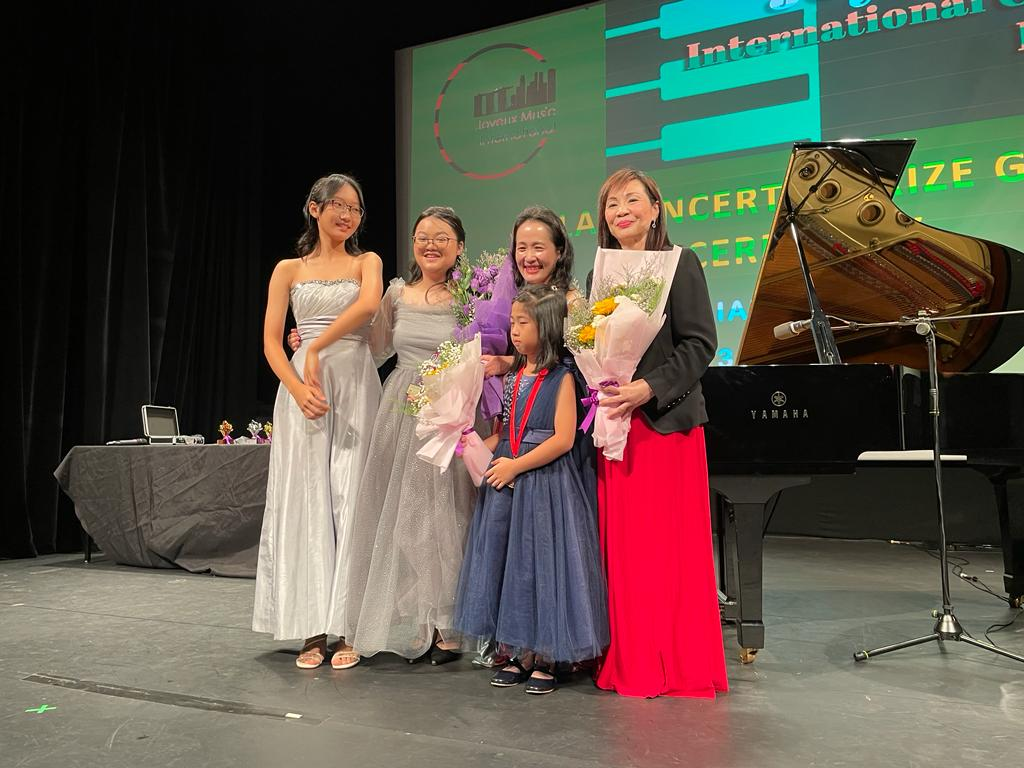 A group of five women and one girl standing on stage, with a grand piano to their right and a large screen behind them displaying text related to music and international awards. Some women are holding bouquets of flowers, and all are smiling.