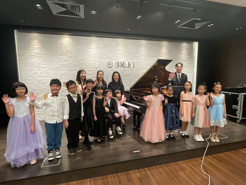 Group of children and an instructor posing on a stage with a Yamaha piano, some children in formal dresses and others in casual clothes, in a music or recital setting.