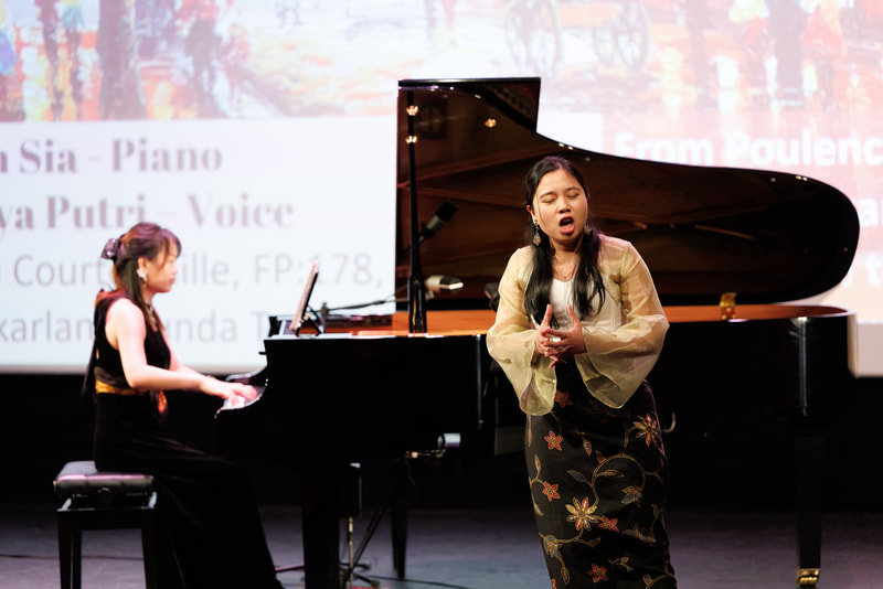 A young woman sings while another woman plays the piano on stage during a performance.