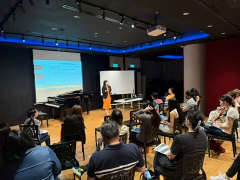 A classroom with a teacher at the front, students seated in chairs, some taking notes, and a large screen displaying educational content.