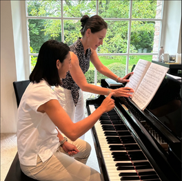 Two girls practicing piano together in a well-lit room with large windows and green trees outside.