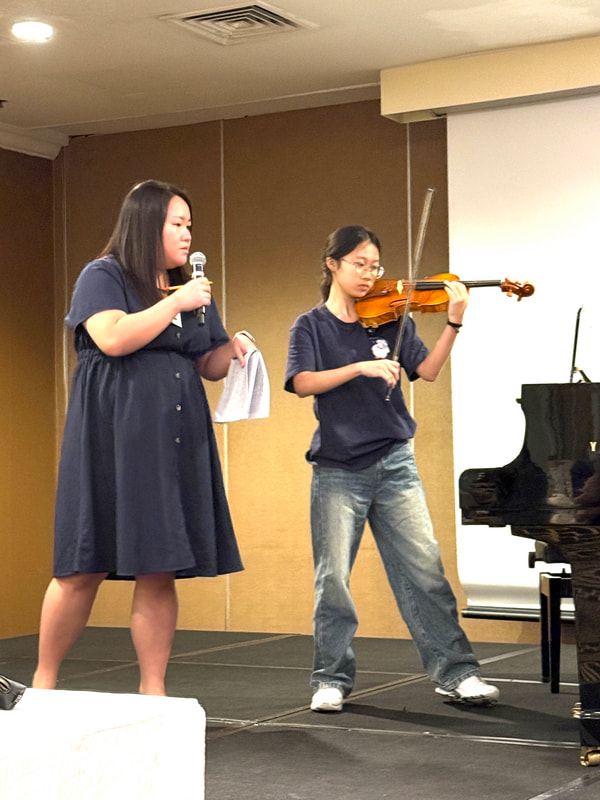 Two young women performing on stage; one singing into a microphone, the other playing the violin.