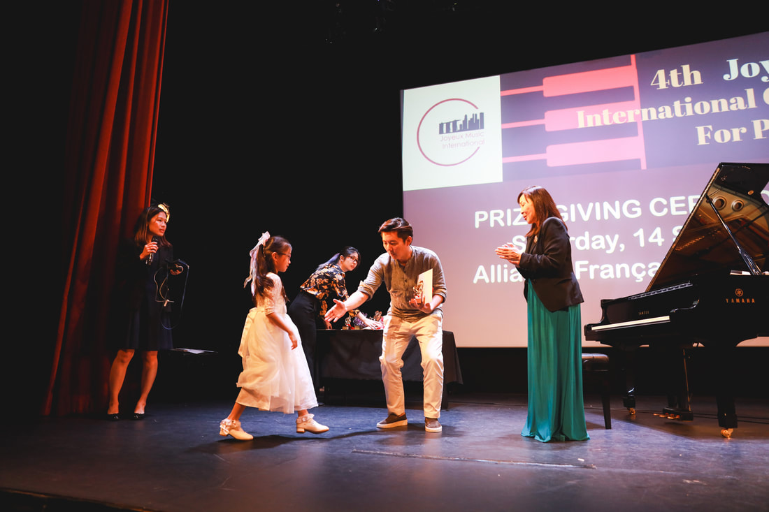 People on stage during an award ceremony, with a large screen in the background displaying event details. One young girl in a white dress is receiving an award from a man, while others observe, and a grand piano is on the right.