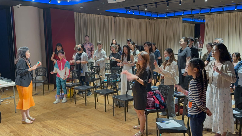 A woman leading a singing or speaking activity with children and adults in a room with chairs and curtains.