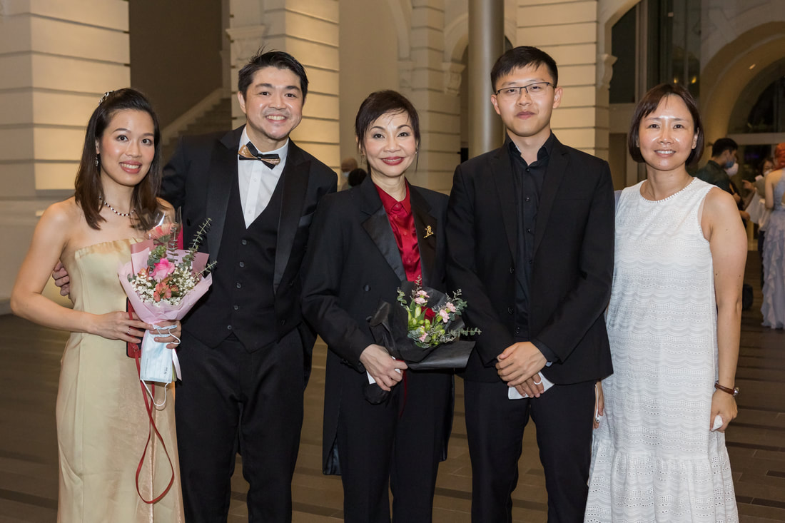 Group of five people dressed in formal attire standing together inside a building, with some holding bouquets of flowers.