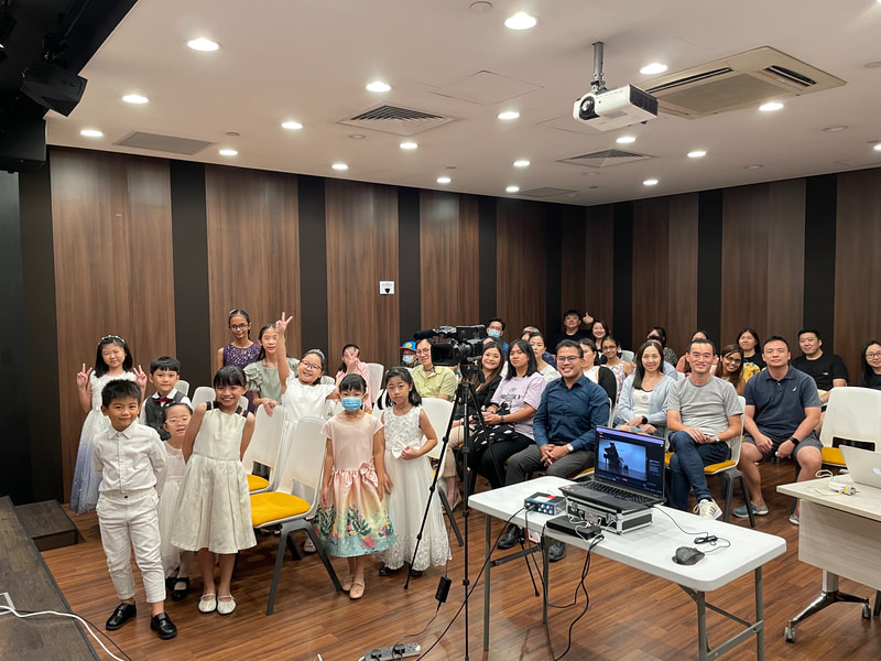 A group of children and adults gathered in a room with wood-paneled walls, some children in dresses and a few wearing masks, seated and standing while smiling and posing for the photo. There is a camera and audiovisual equipment set up in the foregro