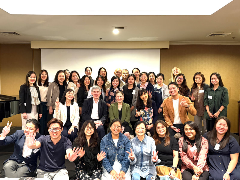 Group of diverse adults posing for a photo in a conference room, some making peace signs, smiling, and dressed in business casual attire.