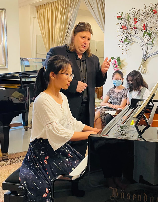 A music teacher instructing a young girl on a keyboard at a piano lesson, with two other students and a piano in the background.
