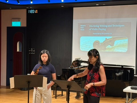 Two young girls standing in front of music stands during a presentation, with a piano and a projected slide titled 'His String Writing and Techniques of Violin Playing' in a music classroom.