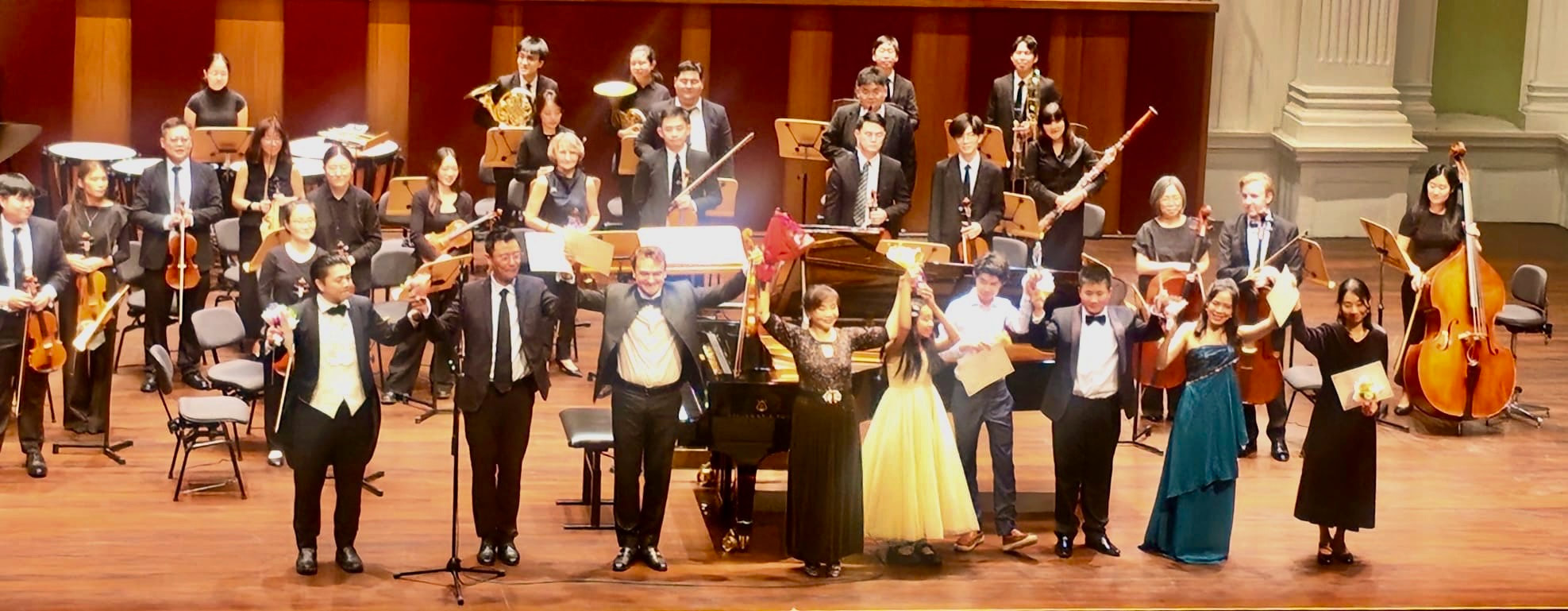 Orchestra group on stage with conductor and soloists holding flowers after a performance.
