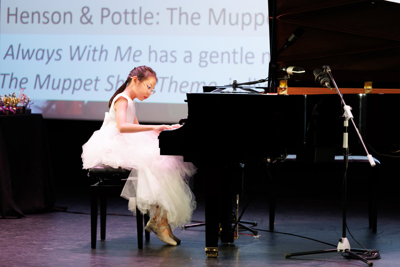 A young girl in a white dress playing grand piano on stage during performance.