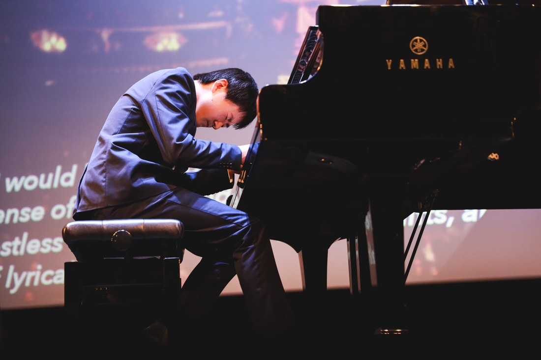 A man in a suit playing a Yamaha grand piano on stage.