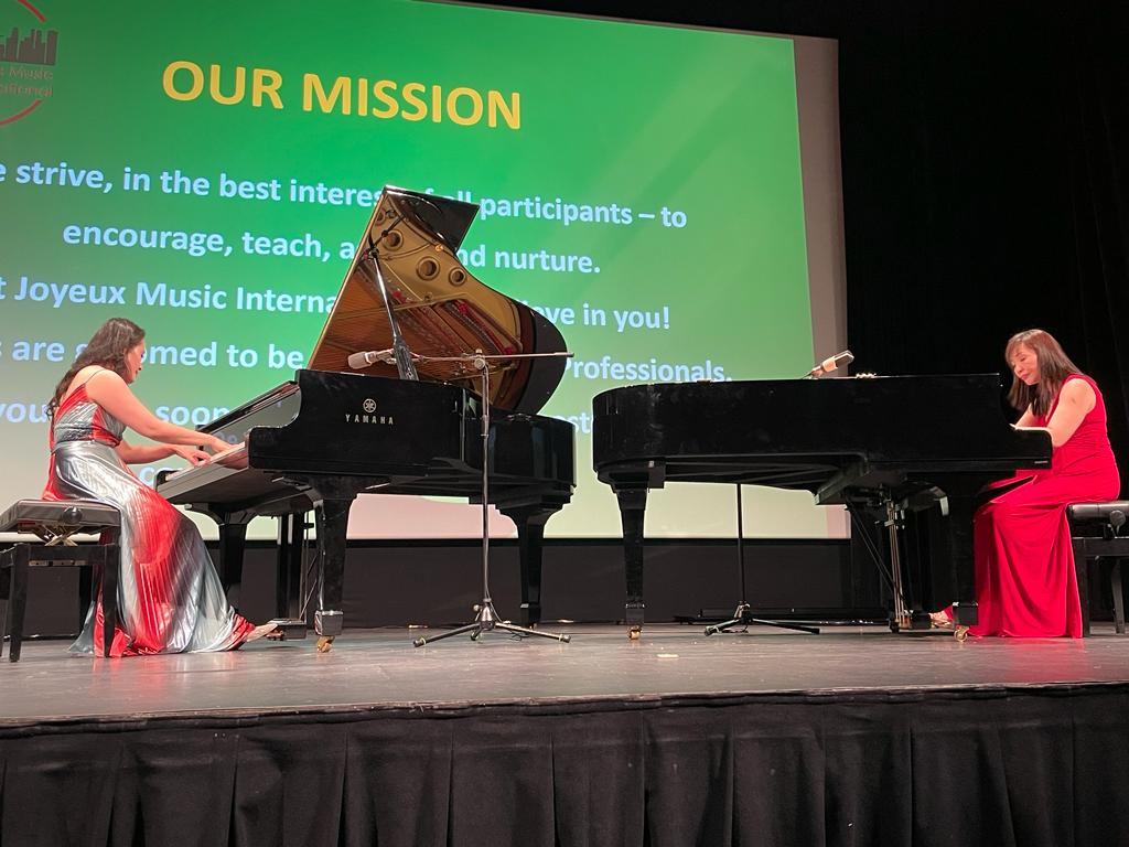 Two women in formal dresses playing pianos on a stage during a concert, with a large screen displaying "Our Mission" and mission statement in the background.