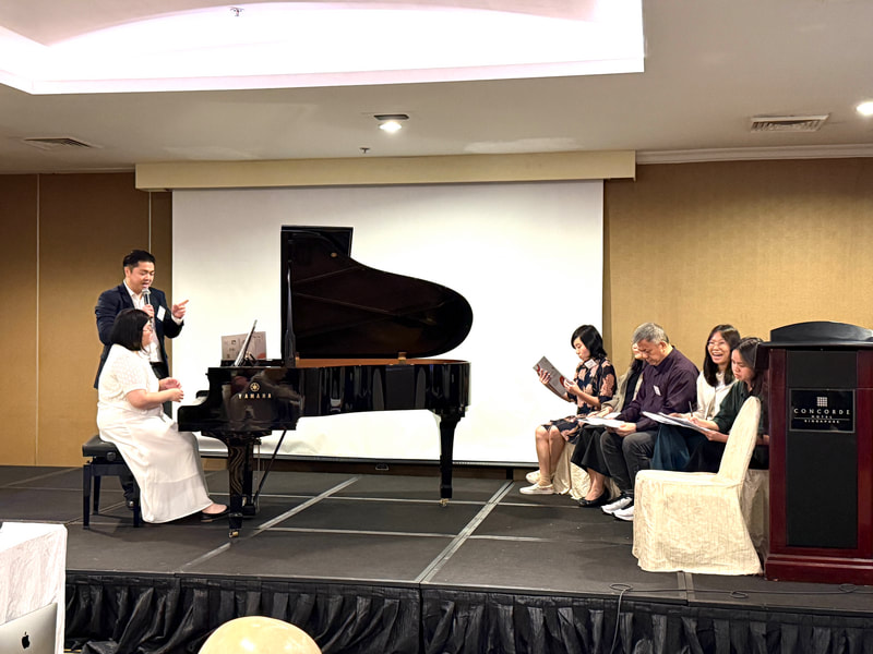 A man in a suit is speaking into a microphone while a woman in a white dress plays the piano on a small stage. Four people are seated to the right, reading sheet music or watching the performance. The setting appears to be an indoor event or presentation with a large white screen behind the stage.