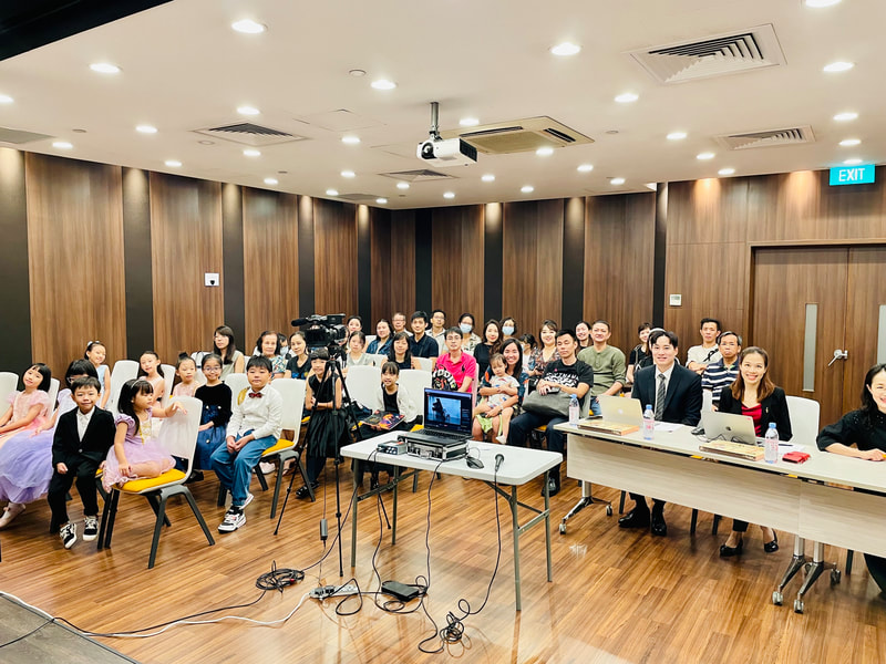 Group of children and adults seated in a conference room, some dressed in formal attire, with laptops, cameras, and a projector, indicating a presentation or event.