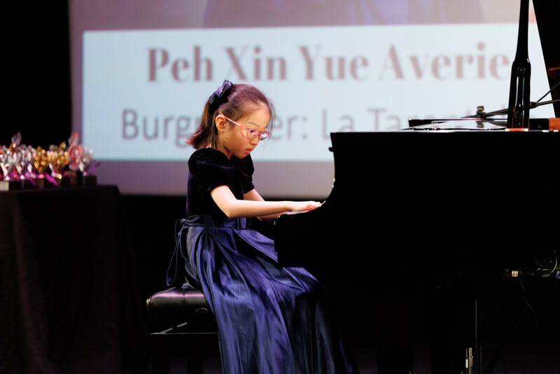 Young girl in a dark blue dress playing the piano on stage, with a screen behind her displaying her name, Peh Xin Yue Averie, and the words 'Burger: La Tarte.'