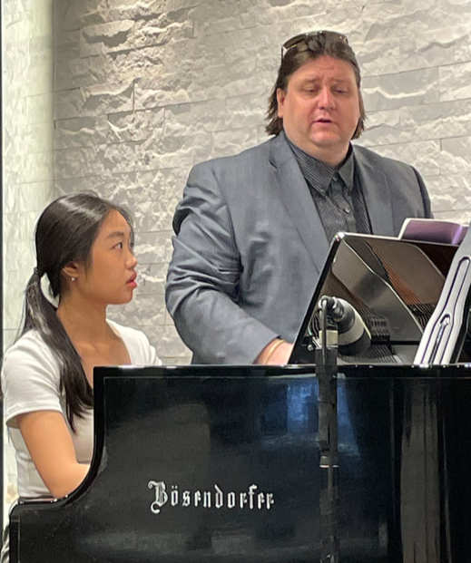 A woman and a man at a grand piano, with the woman playing and the man standing beside and looking at sheet music, in a room with white brick walls.