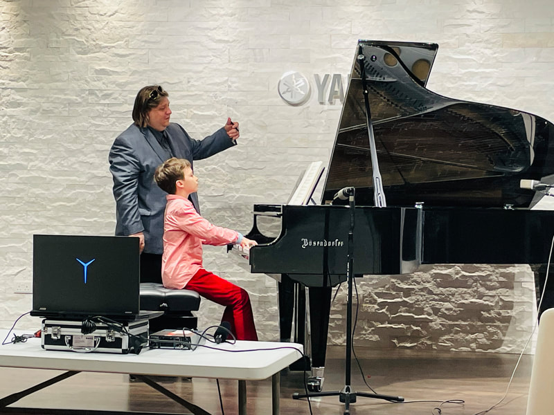 A young boy in a pink shirt and red pants playing a grand piano while a woman in a blazer stands beside him, guiding or instructing, in front of a white brick wall with a Yamaha logo.
