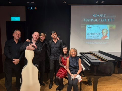 Group of six people posing in a concert hall next to a piano and a mannequin. A screen displays posters for the Mozart Festival Concert.