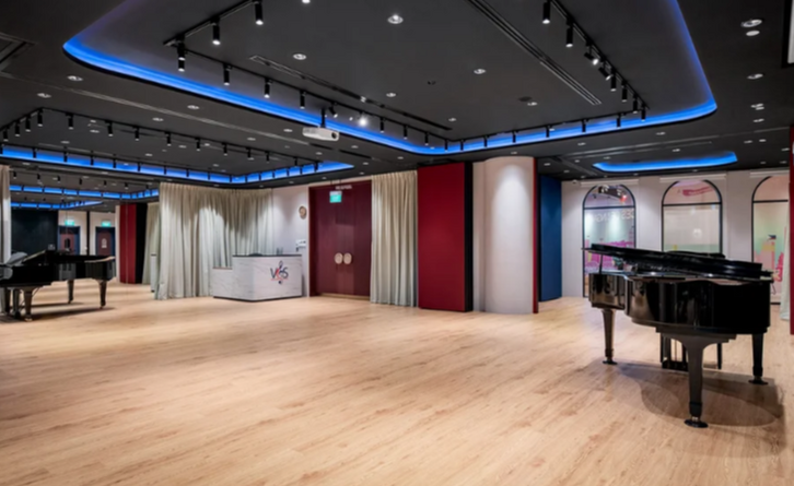 Spacious music studio with grand pianos, modern lighting, a reception area, and a polished wooden floor.